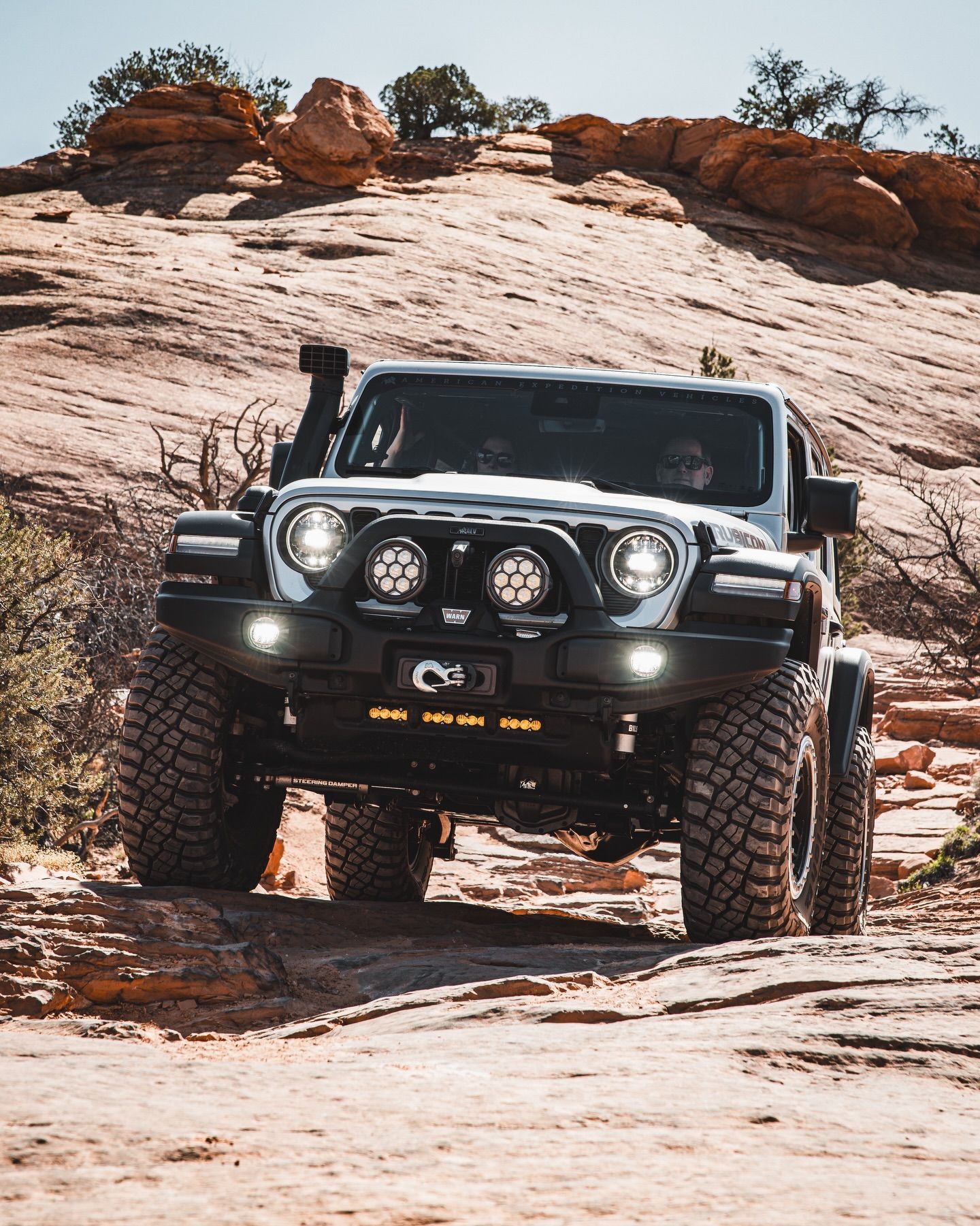 A silver Jeep Wrangler with off-road modifications drives up a rocky, sunlit desert slope.