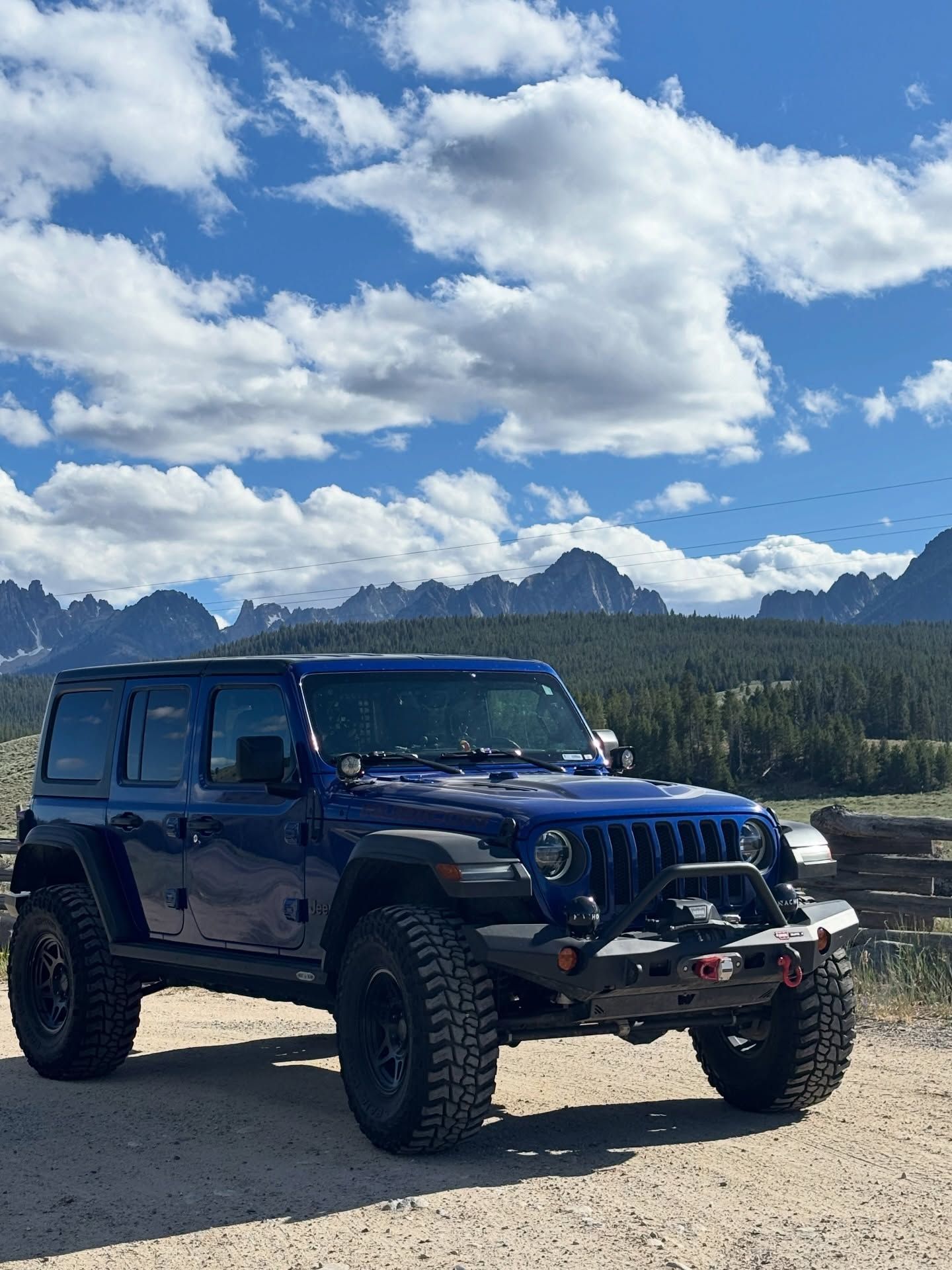 A dark blue Jeep Wrangler parked on a dirt path with rugged, jagged mountains and a cloudy blue sky in the background.