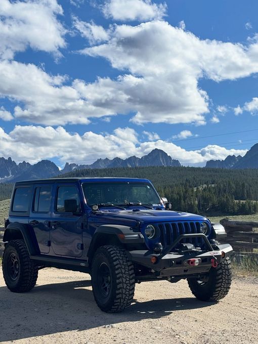 A dark blue Jeep Wrangler parked on a dirt path with rugged, jagged mountains and a cloudy blue sky in the background.