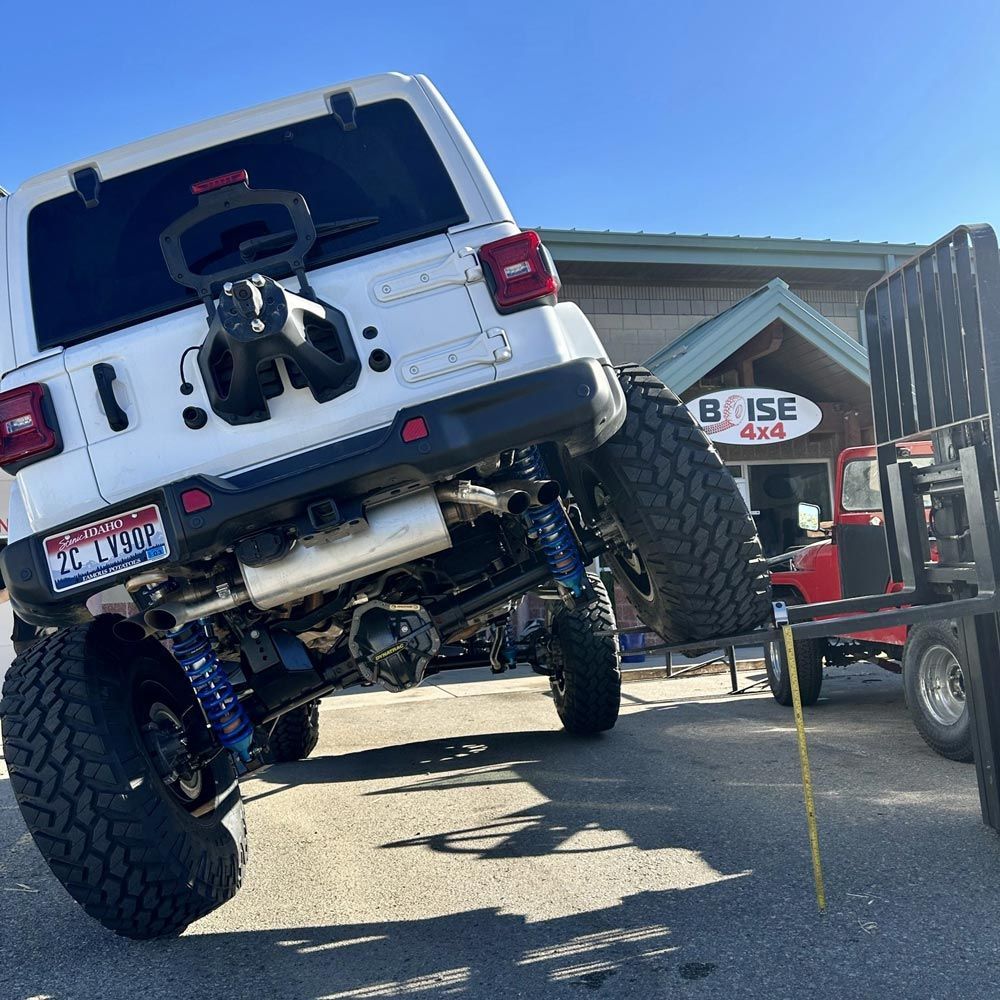 A white Jeep is lifted by a forklift at Boise 4x4, showcasing its extensive suspension articulation and large tires.