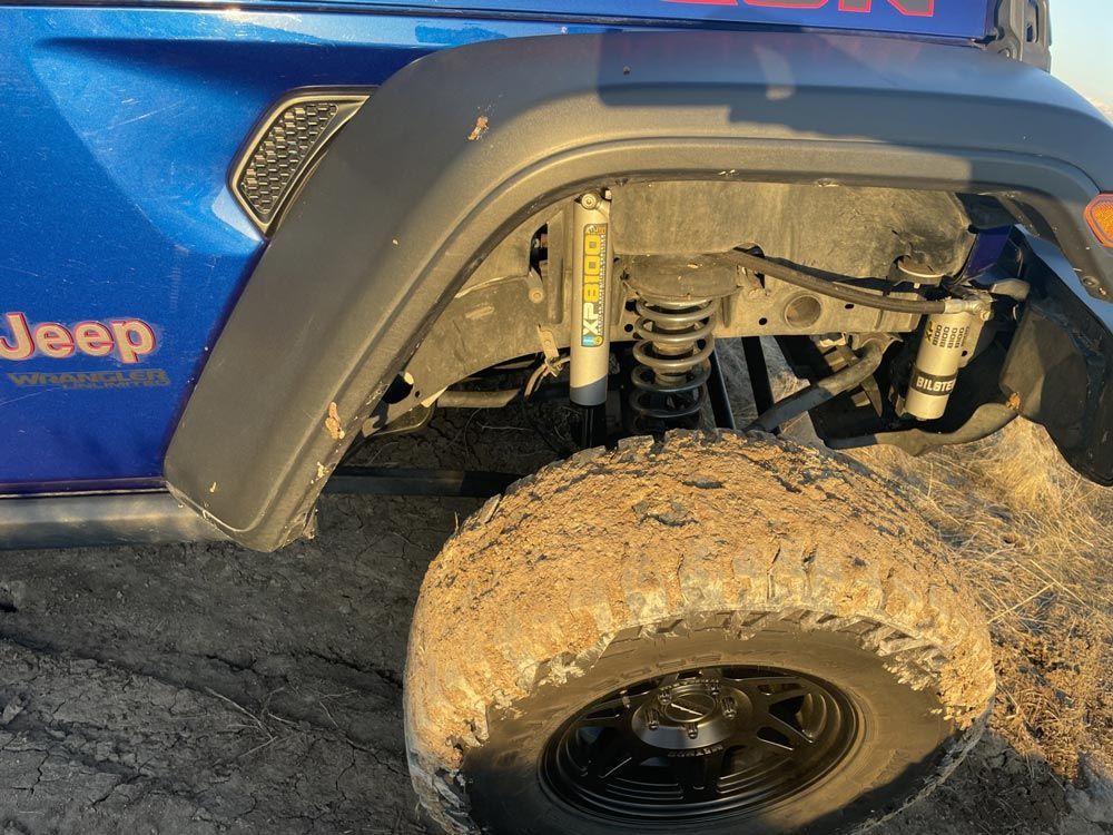 Close-up of a white Jeep Rubicon's front suspension featuring a prominent blue coil-over shock and large off-road tire.