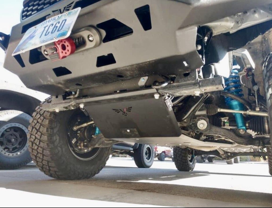 A low-angle view of a truck’s front undercarriage, featuring a dark metal skid plate, winch, and bright blue shock absorbers.