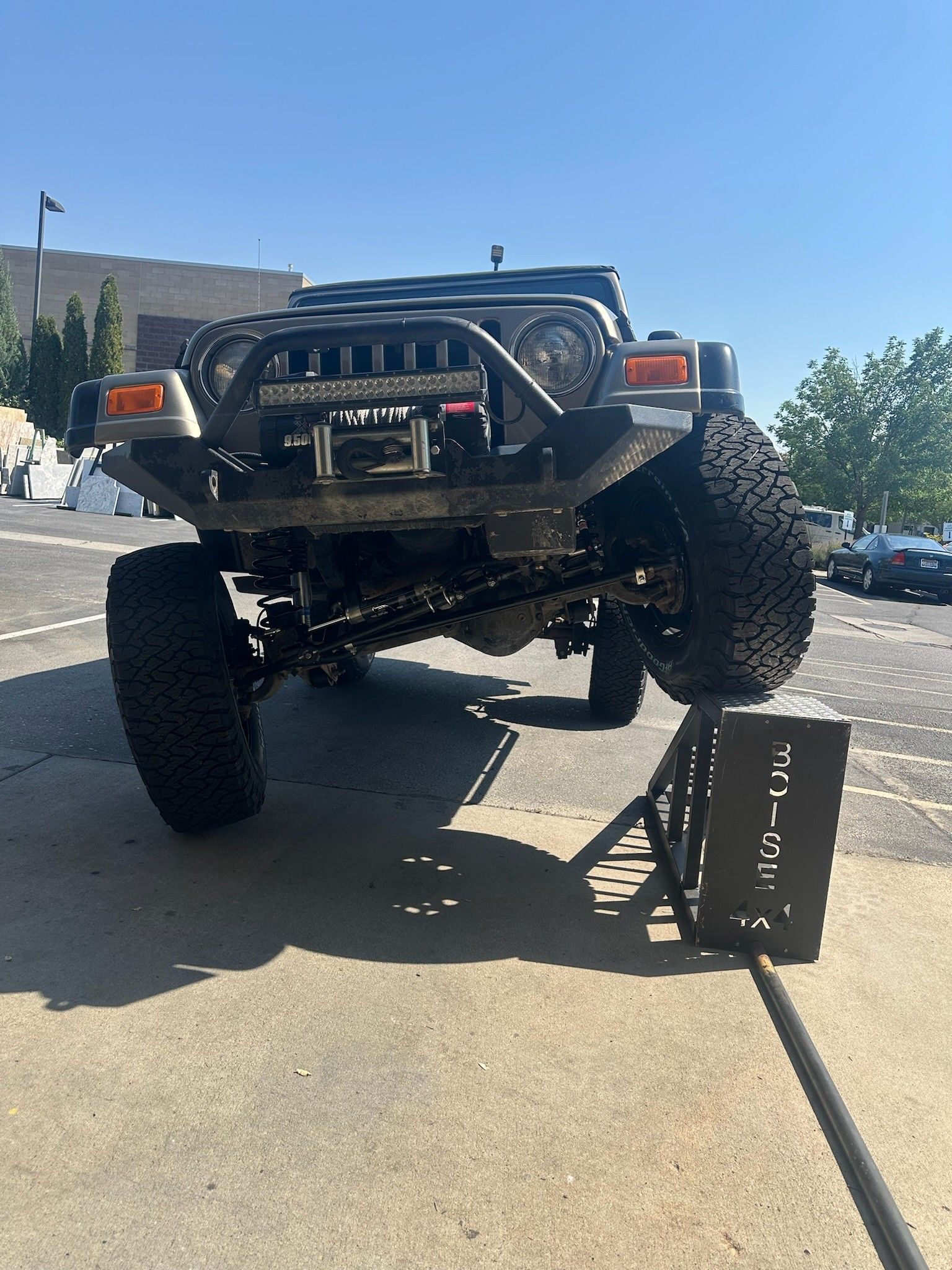 A modified Jeep with large off-road tires parked in a parking lot, with its front driver-side wheel on a metal ramp.