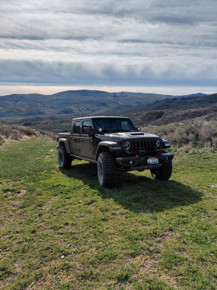 A dark Jeep Gladiator truck parked on a grassy hill overlooking a vast, hilly landscape under a cloudy sky.