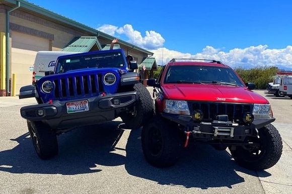 A blue Jeep Wrangler climbs over the front tire of a red Jeep Grand Cherokee in a parking lot.