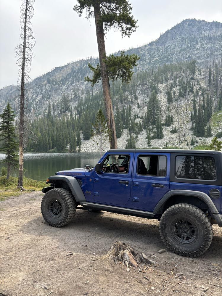 A blue Jeep Wrangler with a black off-road bumper, winch, and rugged tires, parked in front of a forest and lake backdrop.