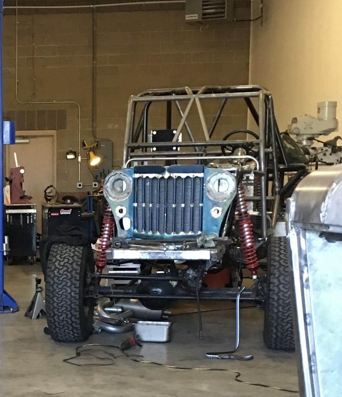 A blue Jeep front end and roll cage under construction inside an industrial workshop with tools and parts visible.