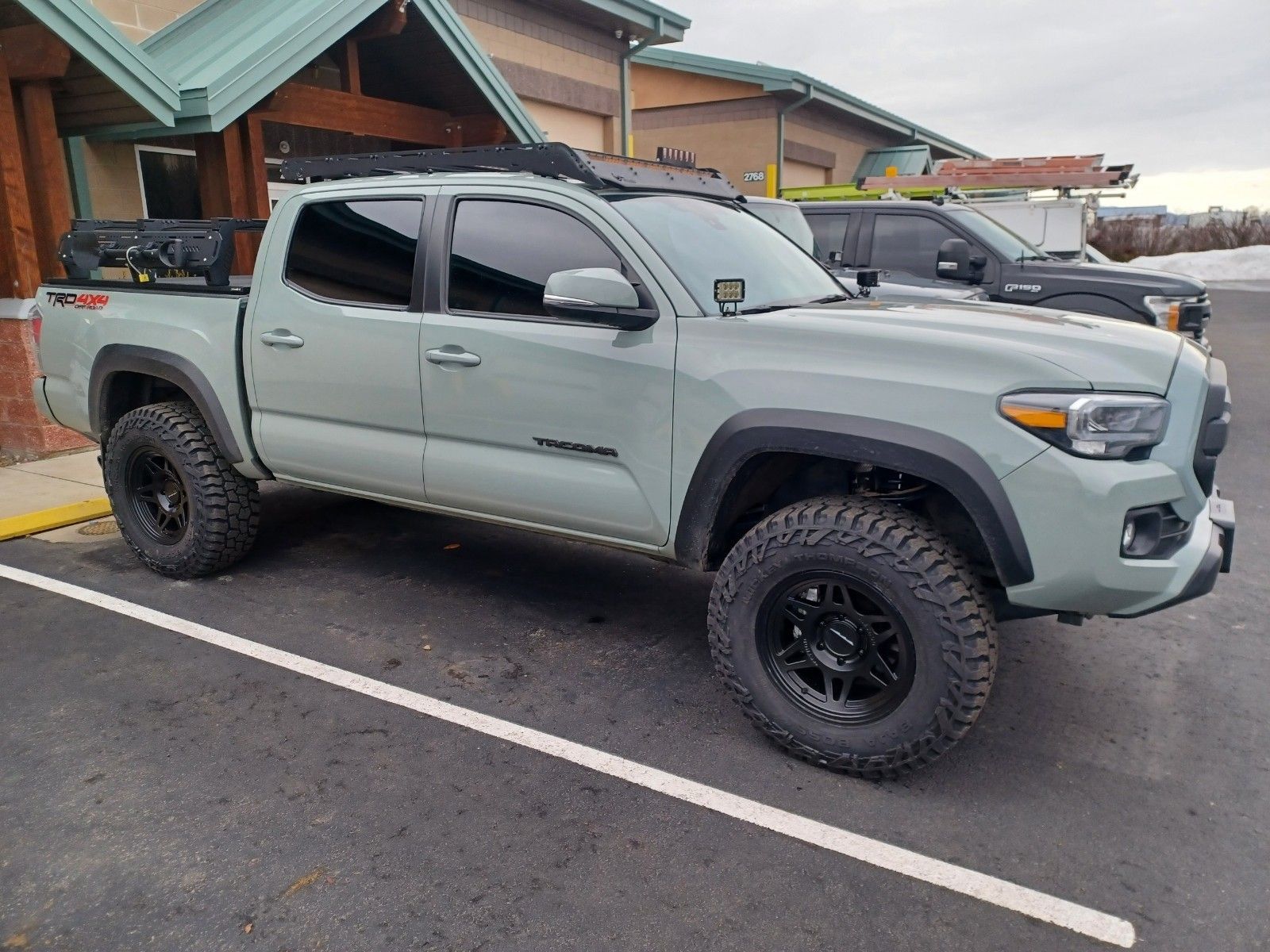 A gray Toyota Tacoma pickup truck with off-road tires, a roof rack, and a bed rack parked in a paved lot.