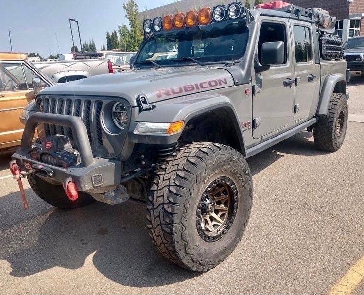 A modified gray Jeep Gladiator Rubicon with large off-road tires, a front winch bumper, and roof-mounted auxiliary lights.