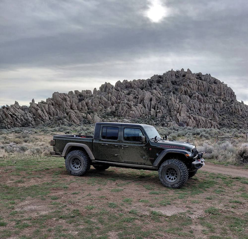 A dark Jeep Gladiator truck parked on a grassy patch in front of a large, jagged rock formation under a cloudy sky.