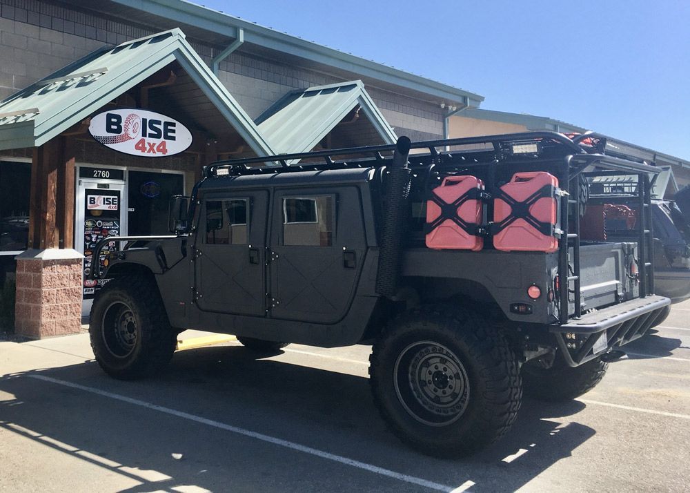 A matte black Humvee with red fuel canisters on the rack parked outside the Boise 4x4 shop on a sunny day.