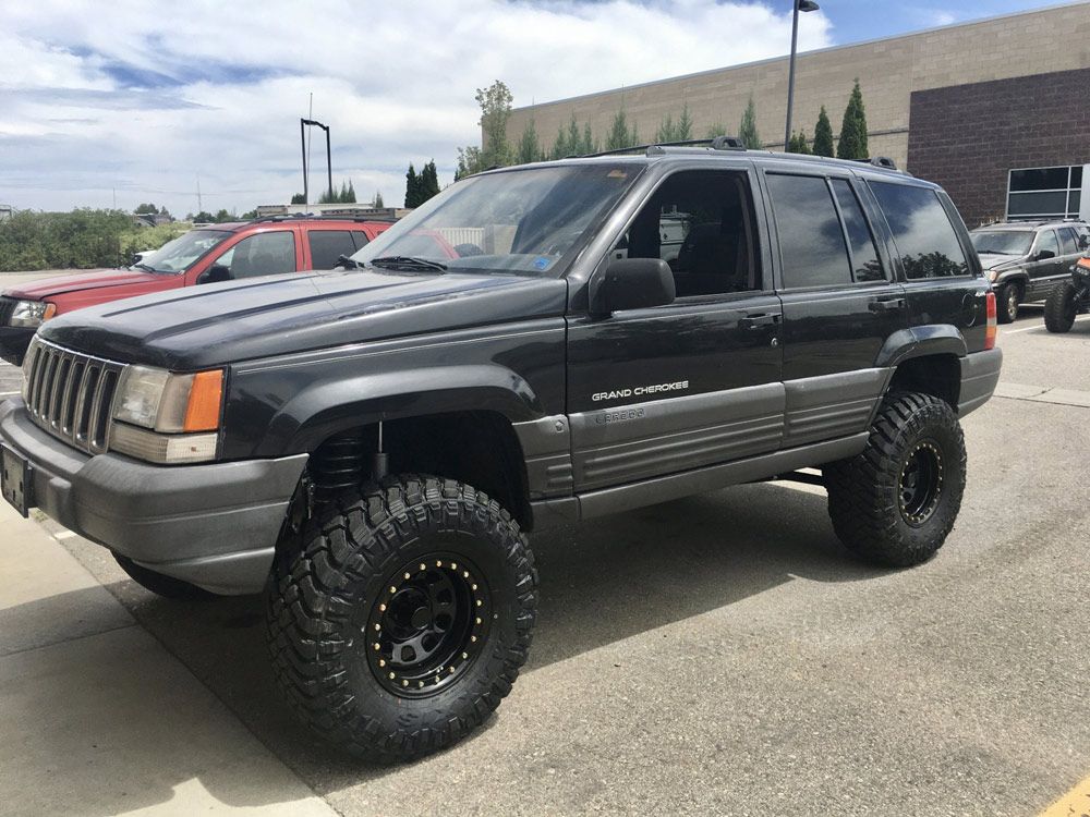 A modified black Jeep Grand Cherokee with oversized off-road tires parked in an outdoor lot on a sunny day.