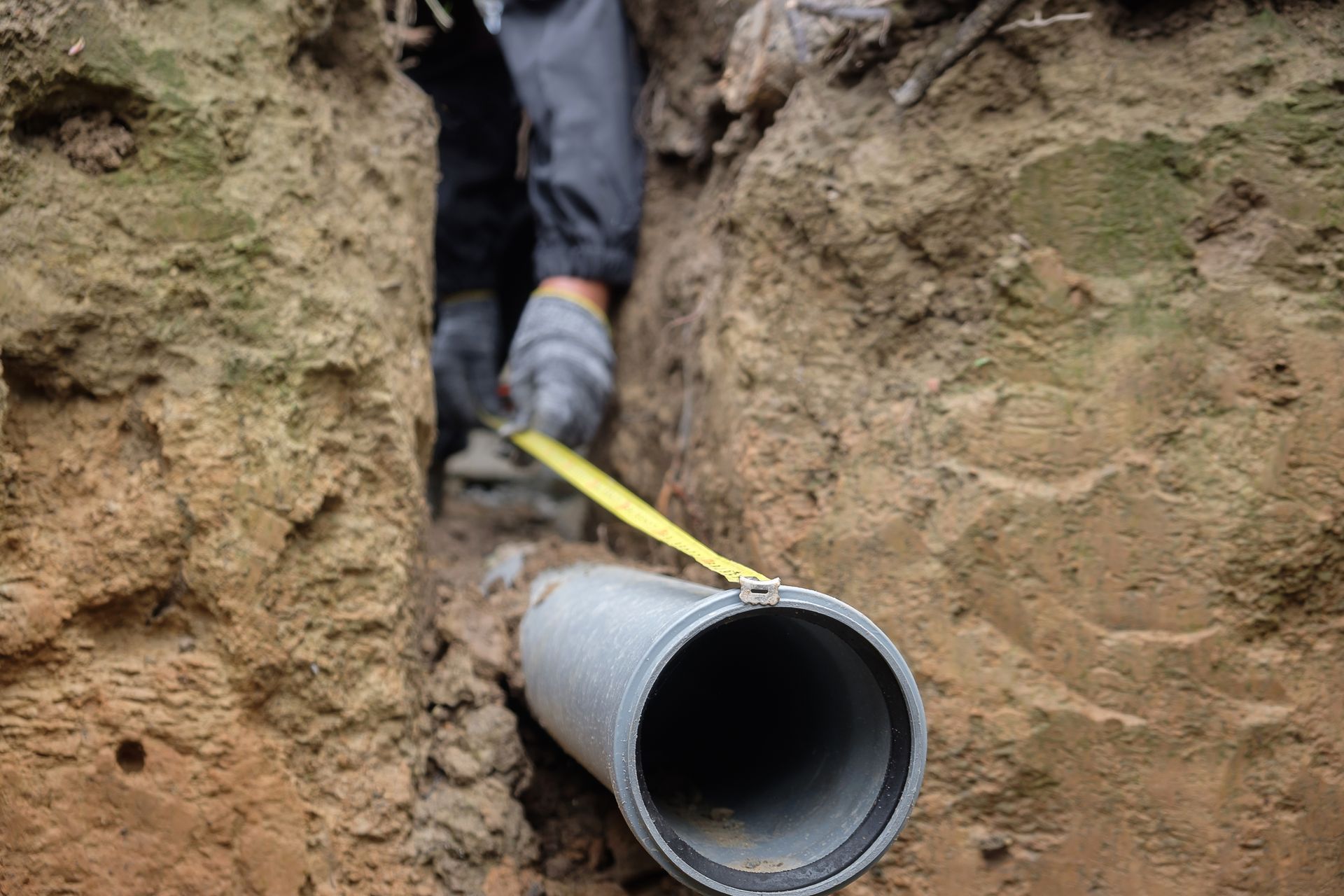 Person measuring a gray pipe in a trench with a yellow tape measure.