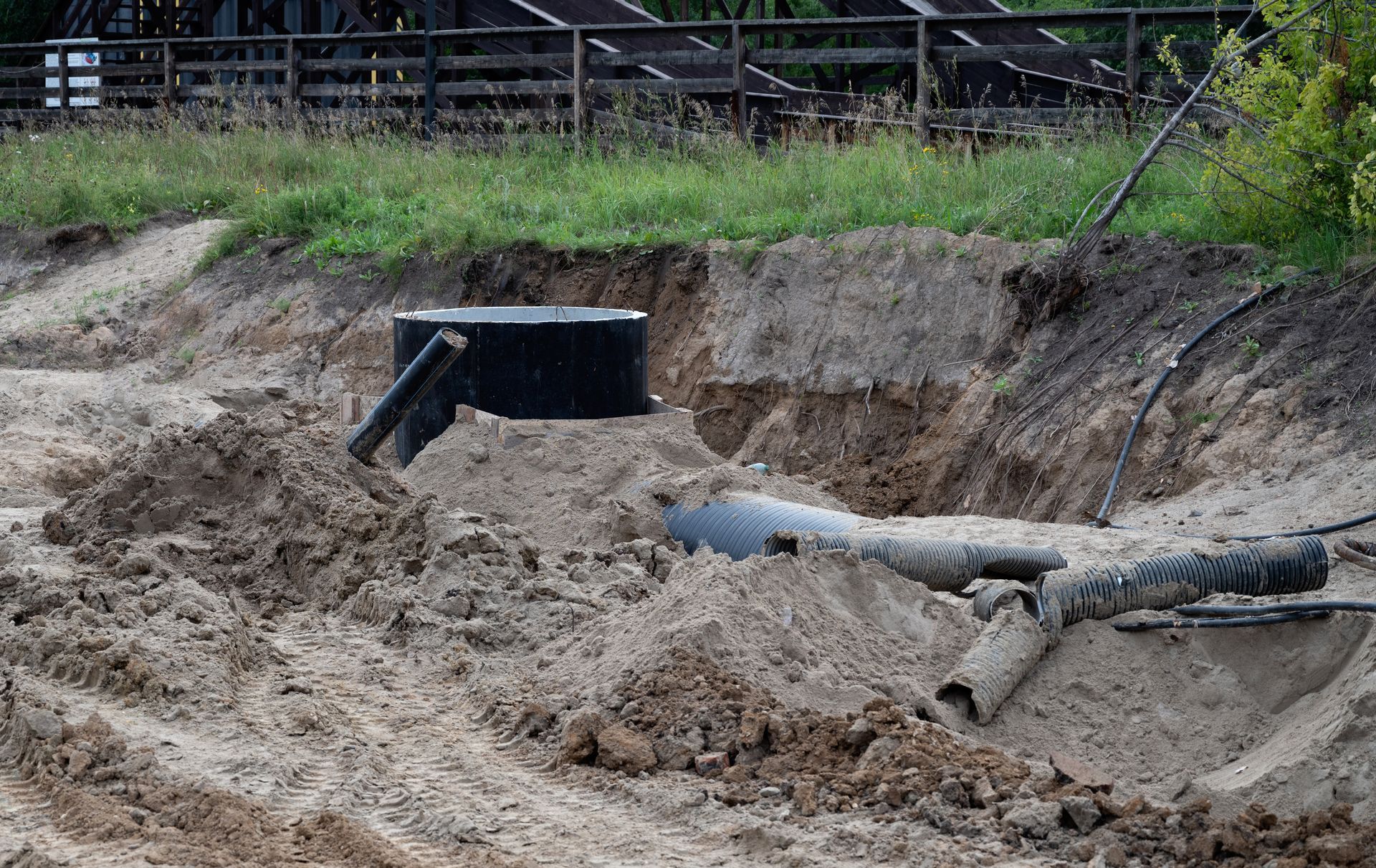 Excavated soil revealing septic tank and pipes with grassy hillside and wooden fence in background.