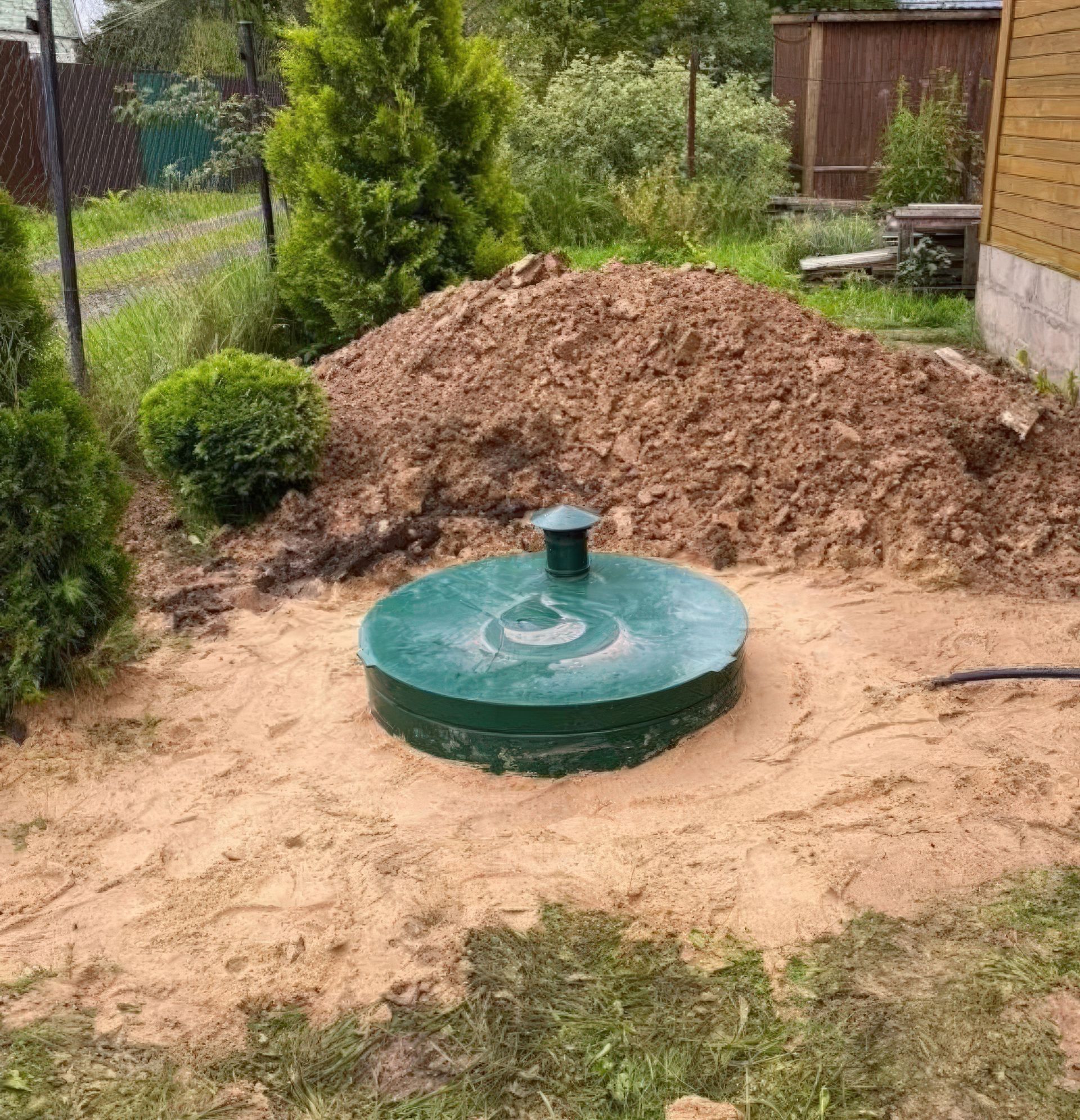 Green septic tank in sandy ground with pile of dirt and vegetation.