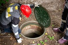 Person in safety gear inspecting an open sewer manhole in a wooded area.