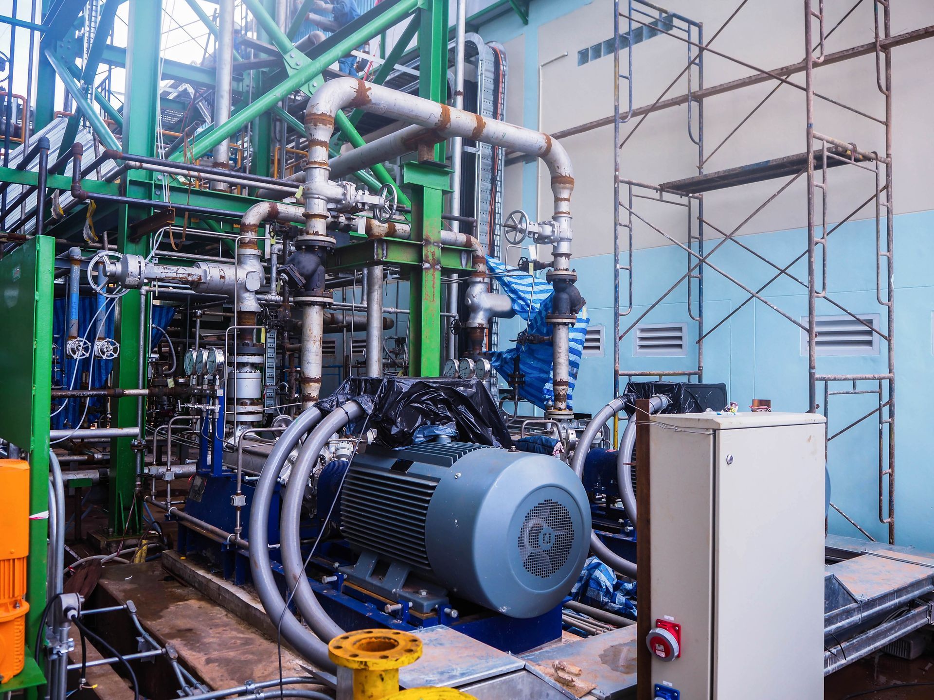 Industrial machinery with pipes and motor in a factory setting, with scaffolding in the background.