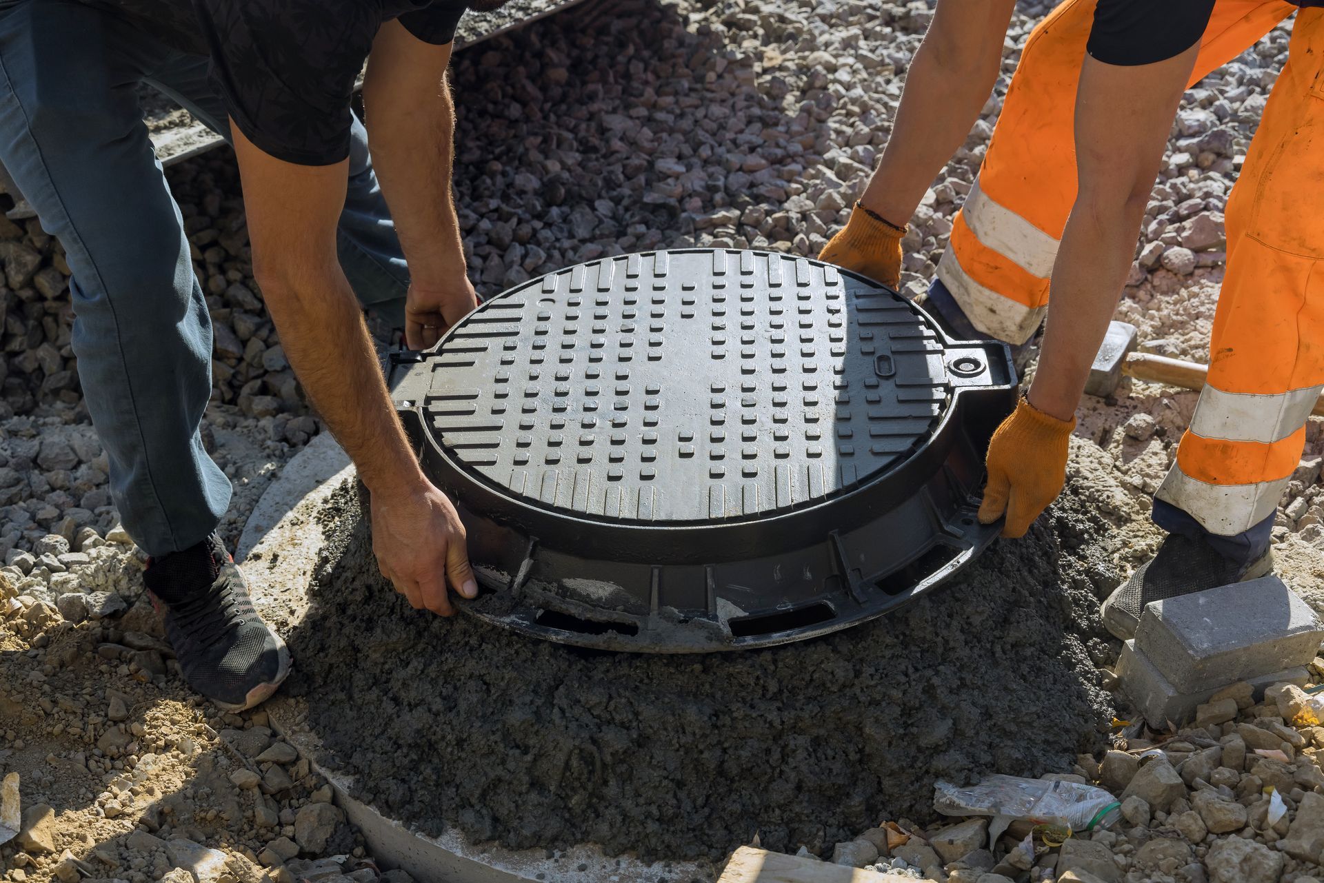 Two workers install a manhole cover in concrete, outdoors. One wears orange pants, the other blue jeans.