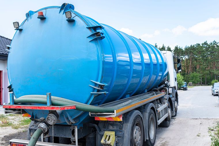 Blue septic tank truck parked on a gravel driveway, hose attached, trees in the background.
