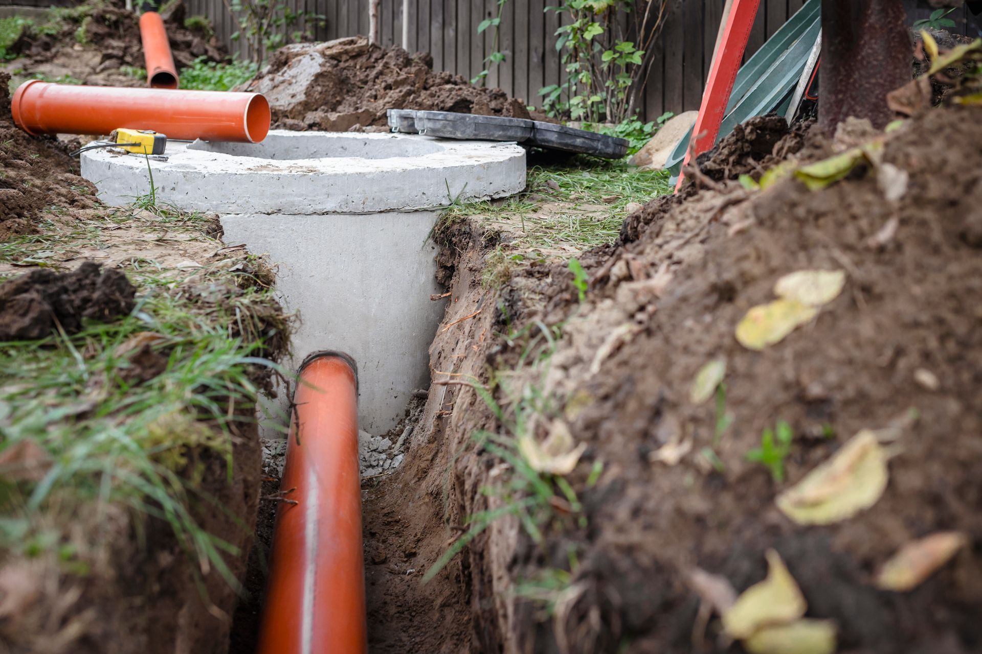Orange drainage pipes connected to a concrete manhole in a dirt trench, outdoors.
