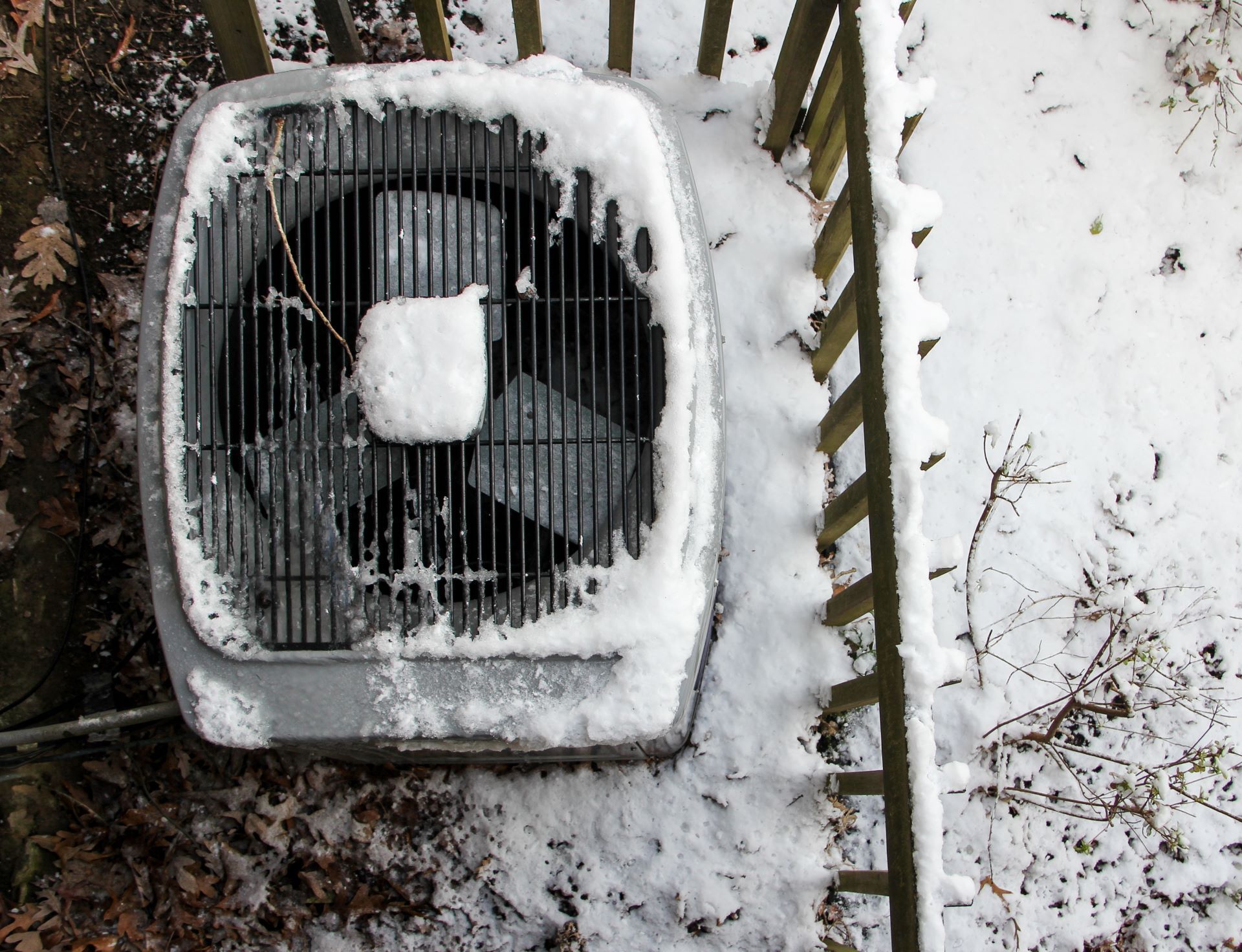 Snow-covered outdoor air conditioning unit next to a wooden fence.