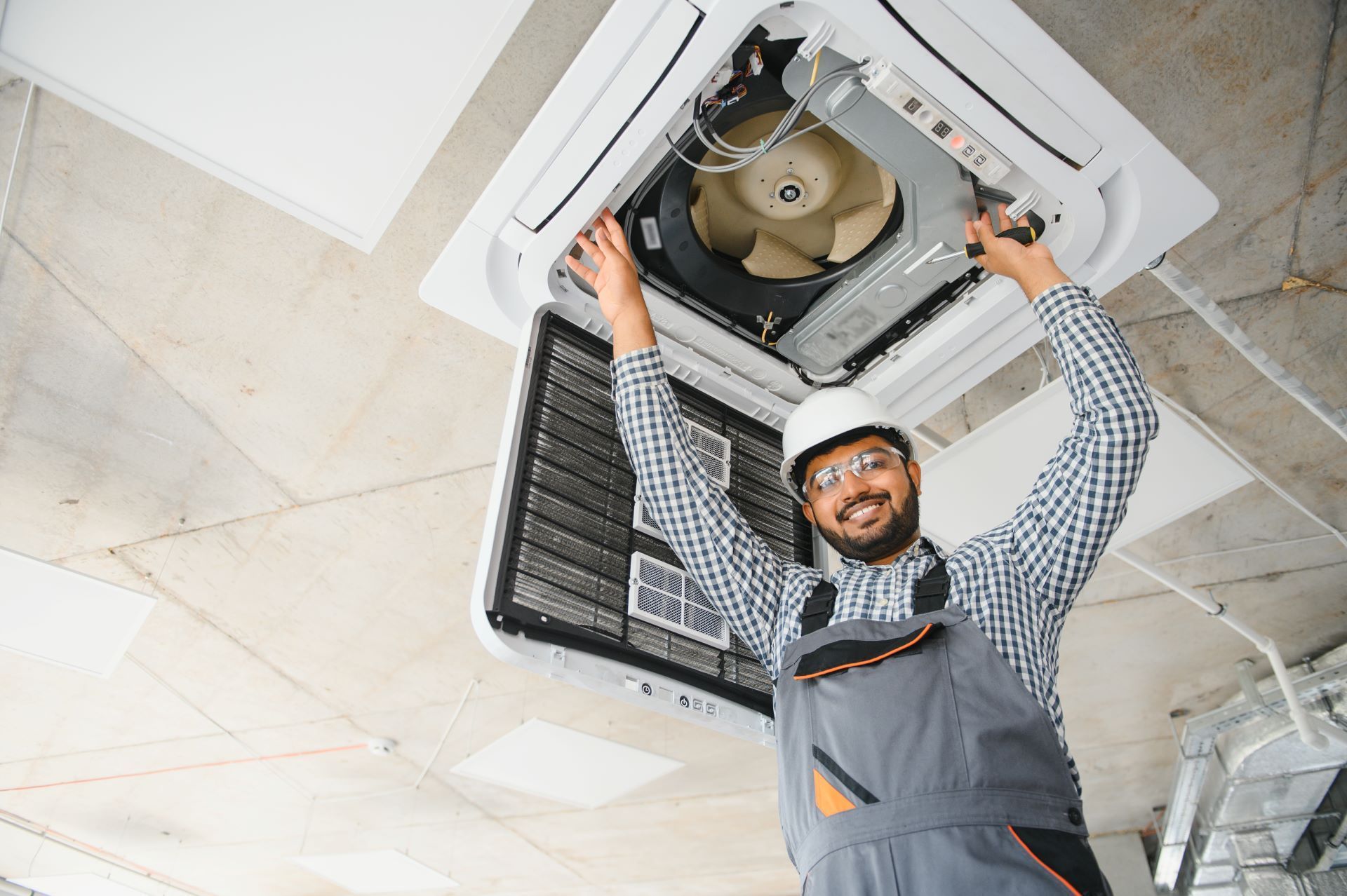 HVAC technician in a white hard hat and goggles works on a ceiling air conditioner.
