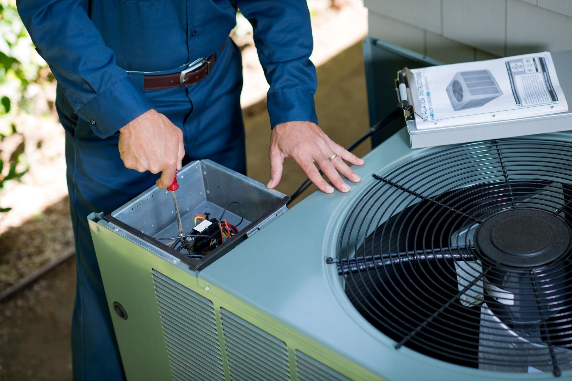 HVAC technician in blue uniform repairs an air conditioning unit outside using a screwdriver.
