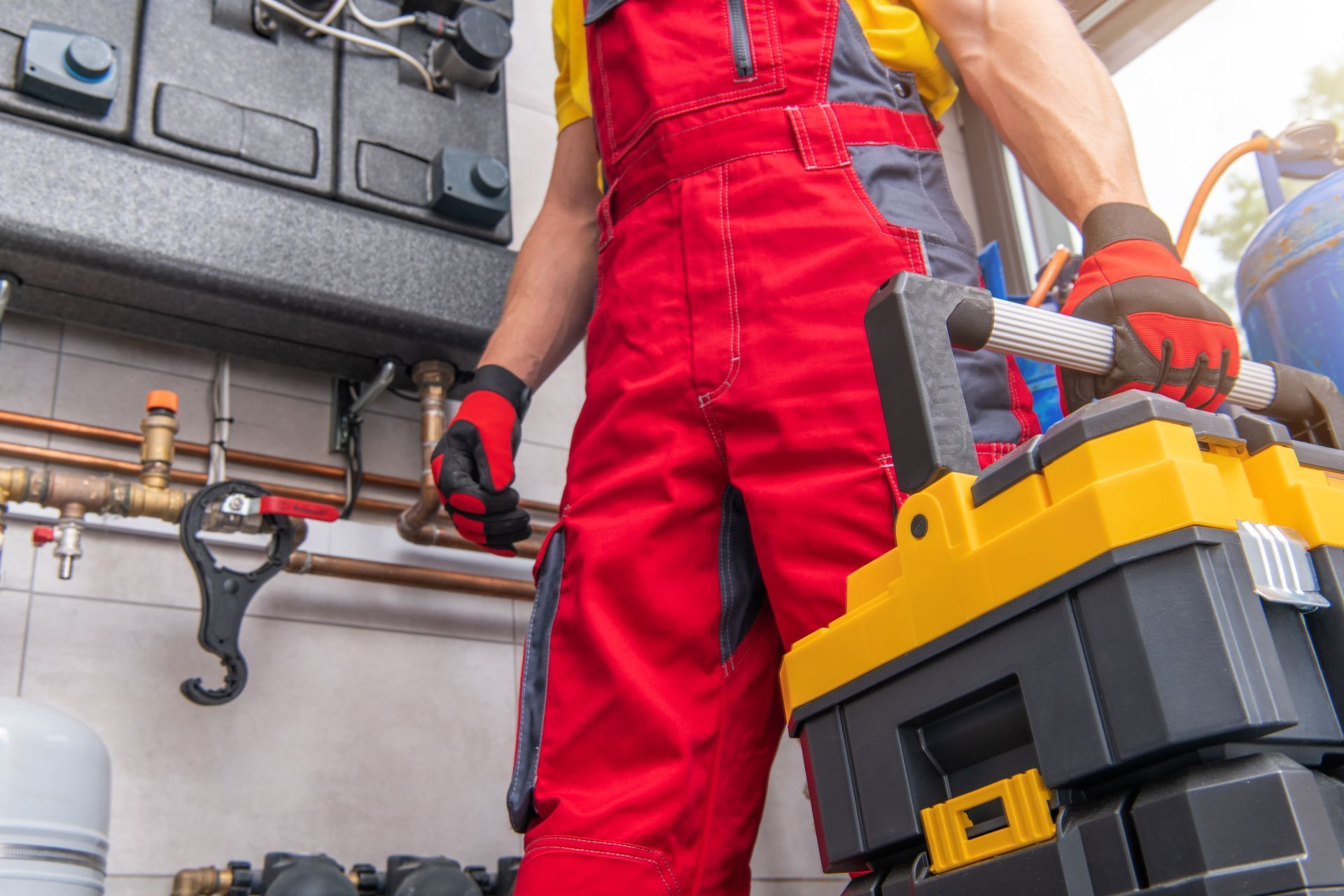 Plumber in red overalls holding a yellow toolbox near pipes and equipment.