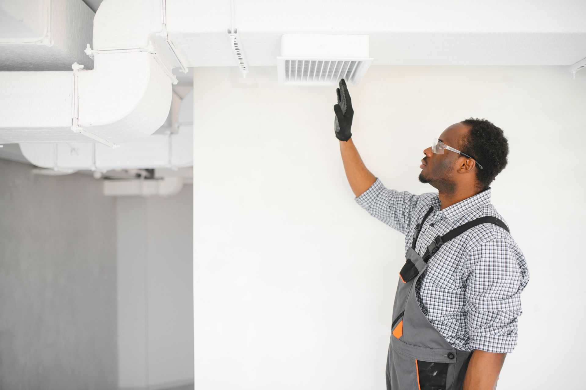 Man in work clothes examines a ceiling vent. White walls, air ducts visible.