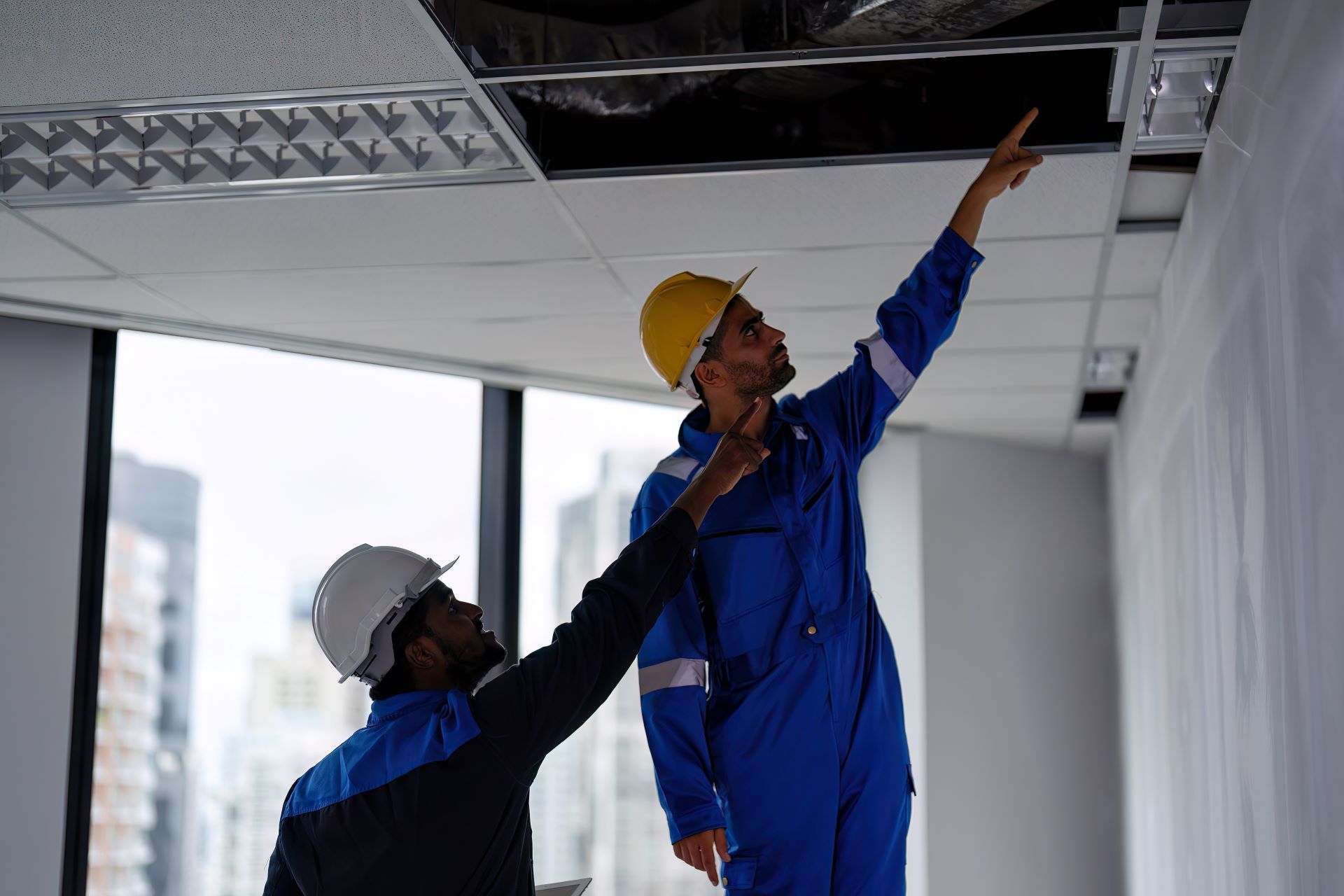 Two construction workers in blue jumpsuits and hardhats inspect a ceiling, pointing up.