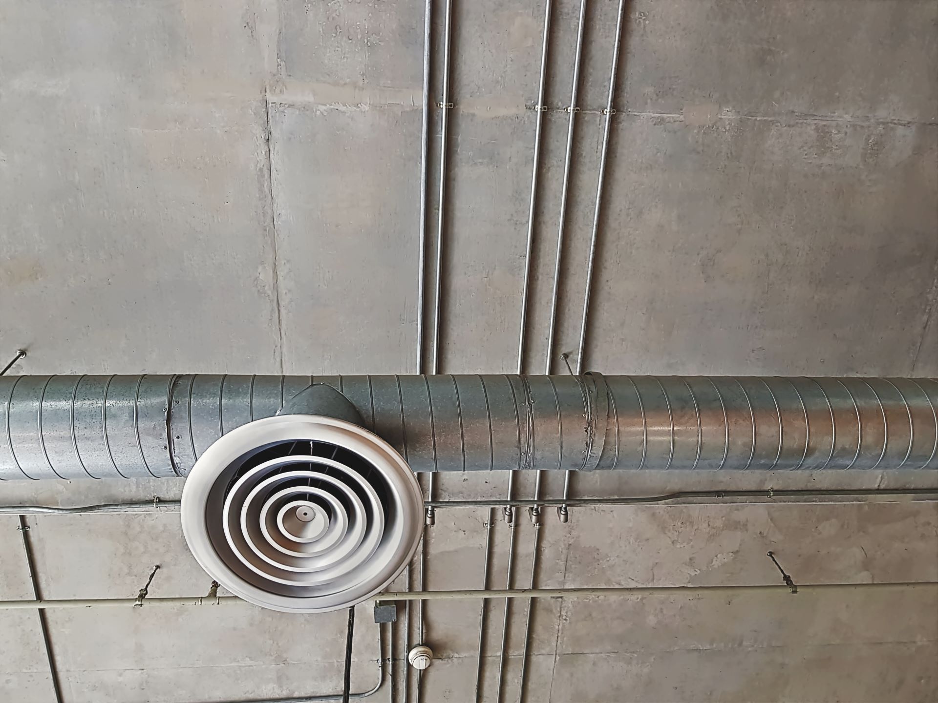 Metal air duct with a vent, electrical conduits, and a gray concrete ceiling.