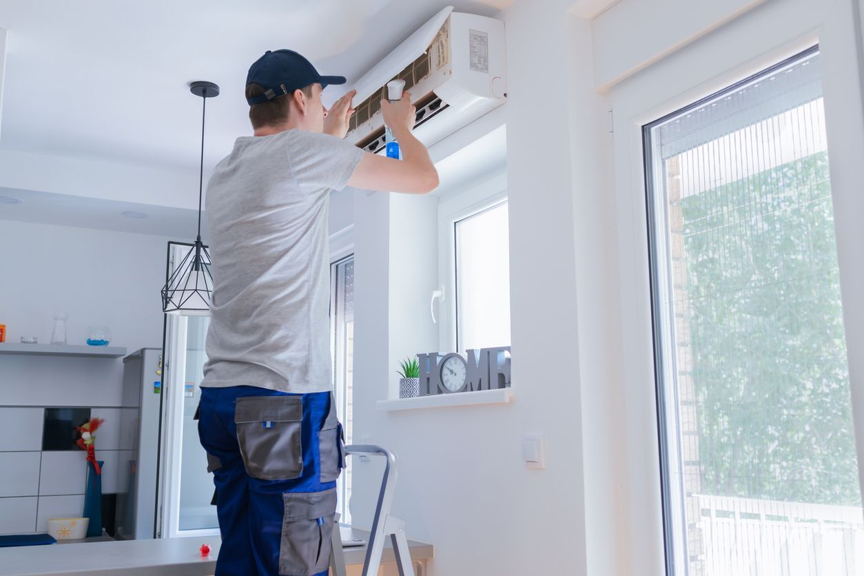 Man in uniform on a ladder installing an air conditioner in a home with windows.
