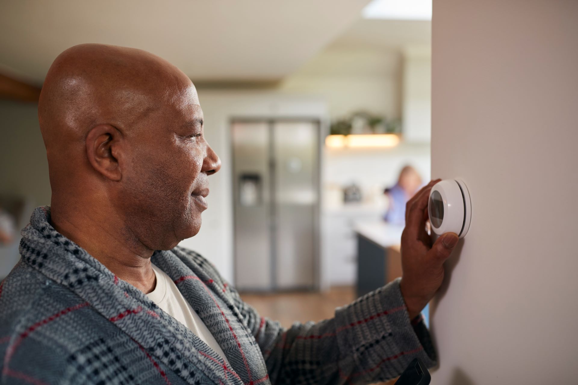 Black man in a robe adjusts a thermostat on a white wall, kitchen in background.