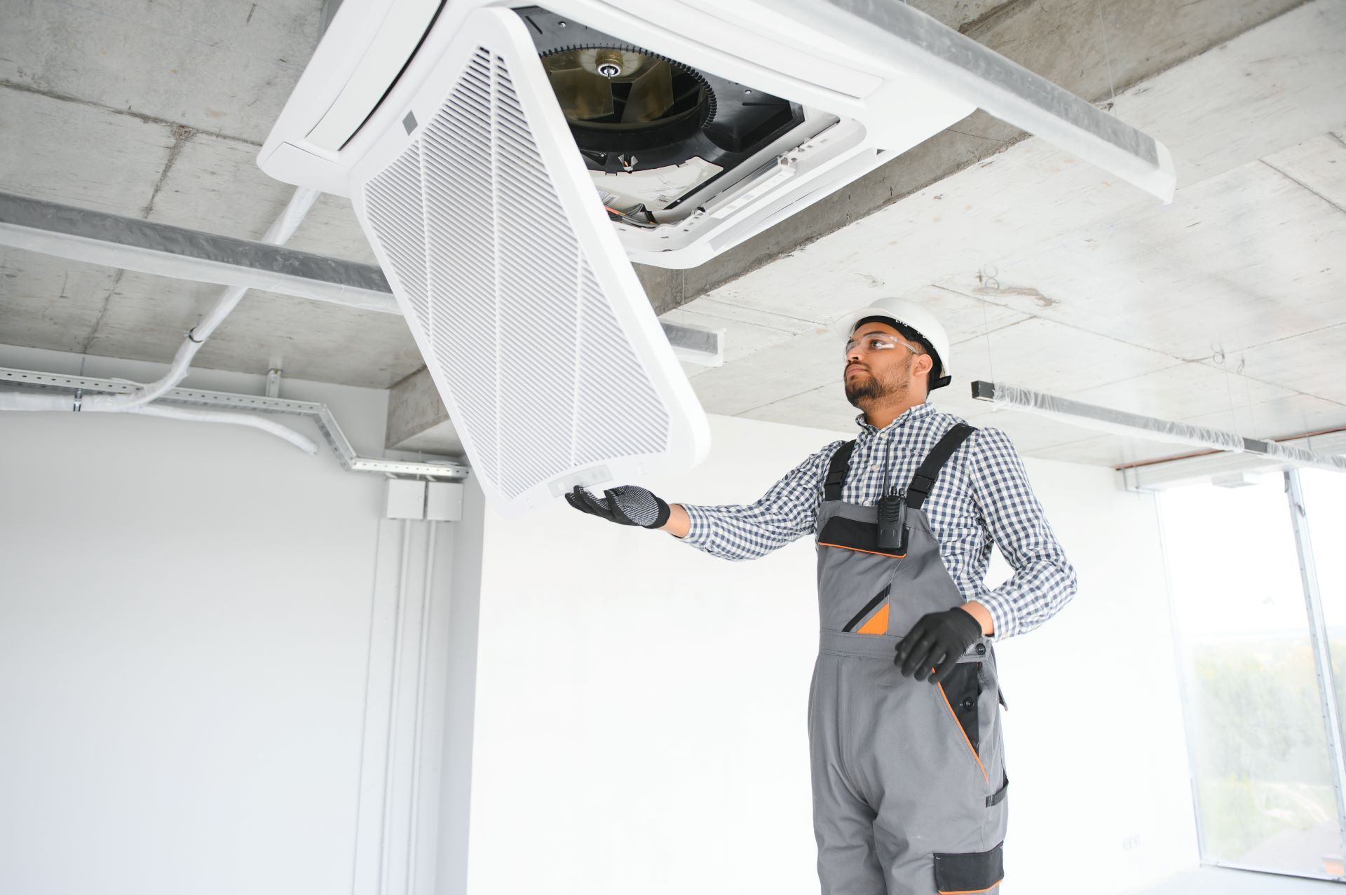 Man in overalls inspecting an air conditioning unit filter in a partially constructed room.