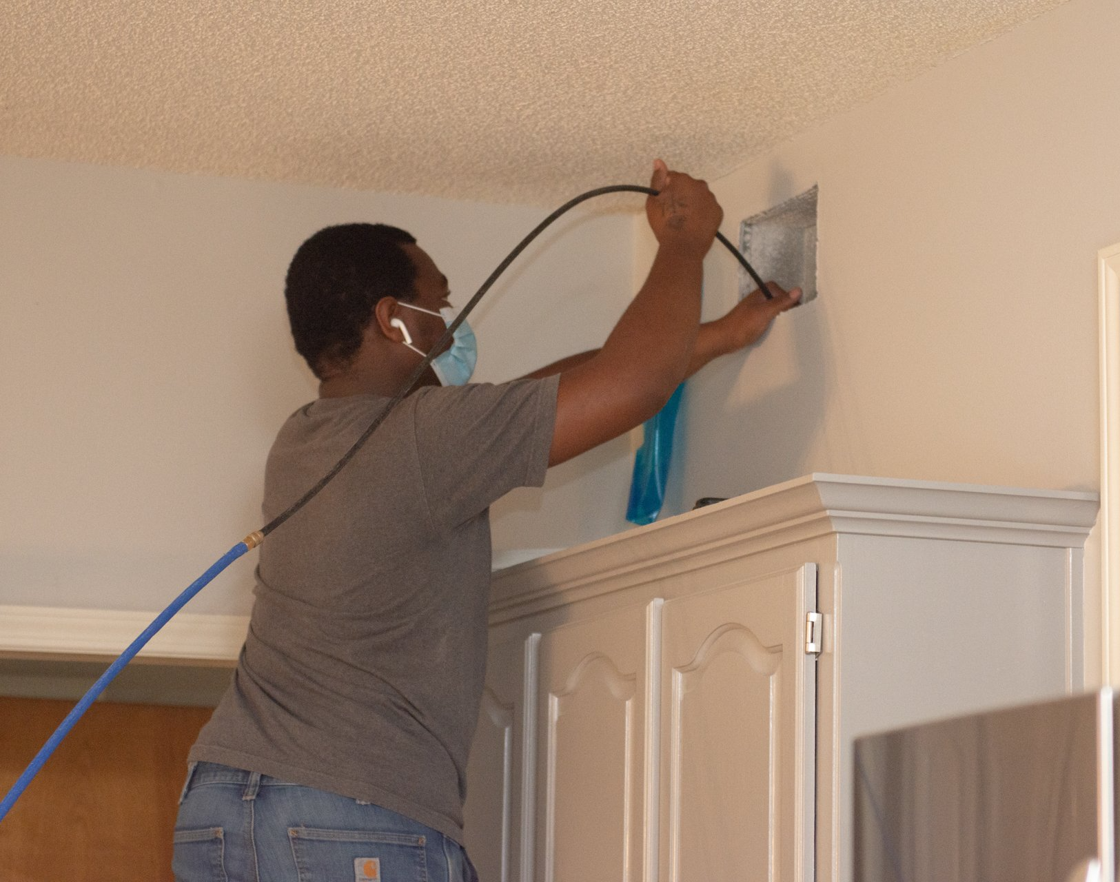 Man cleaning a vent in a kitchen, wearing a mask and using a hose.