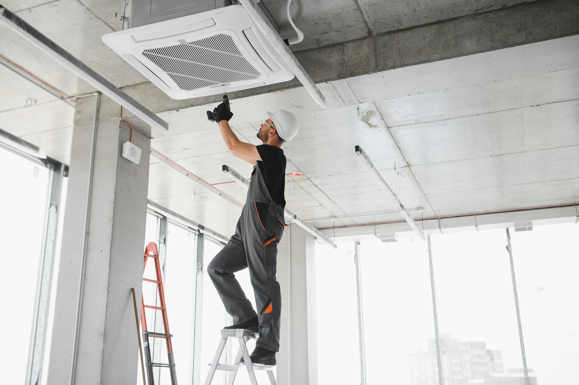 Construction worker in overalls and hardhat on a ladder, installing an air conditioning unit in a building.