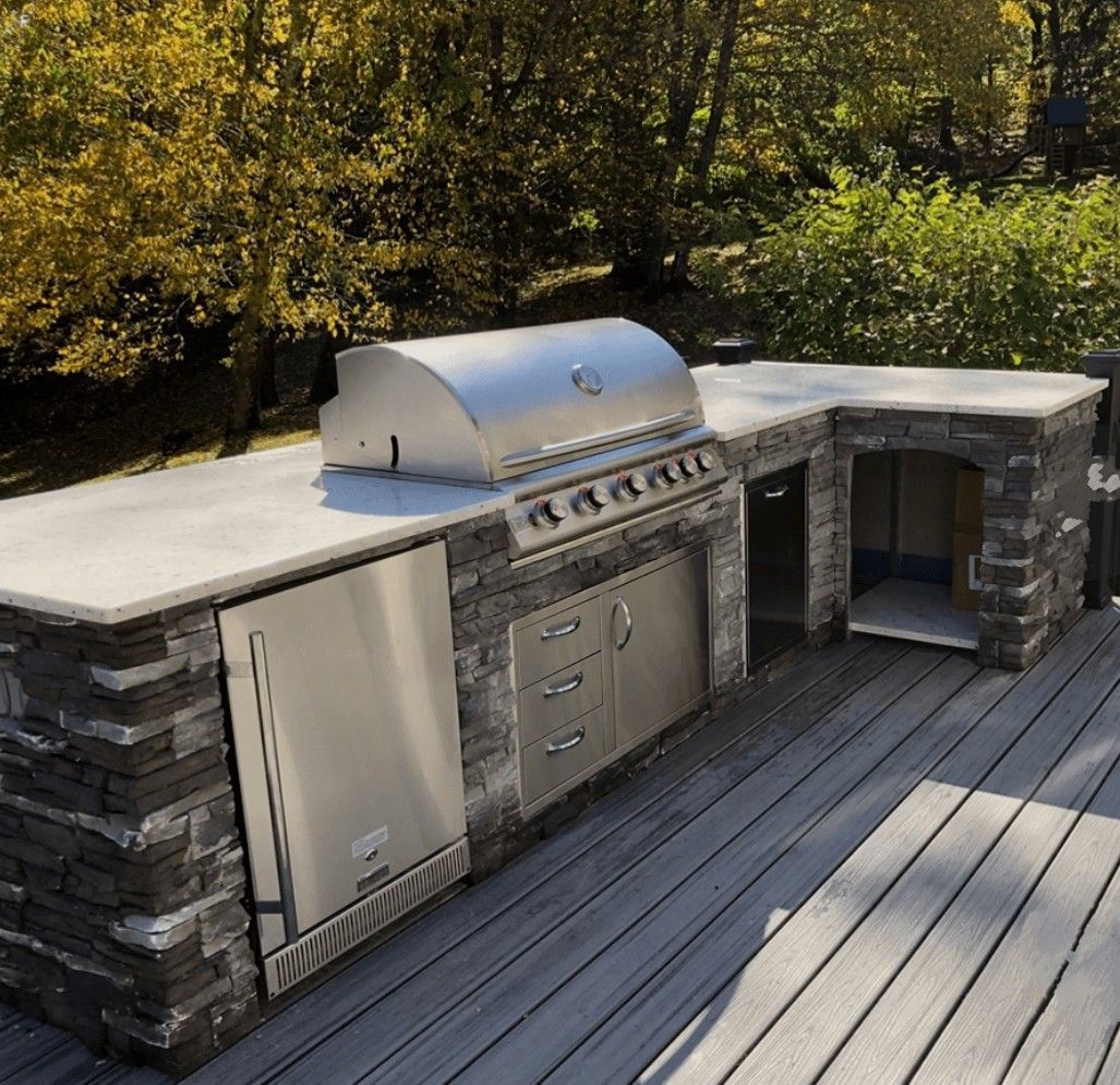 Outdoor stone-grill kitchen with stainless appliances on a wooden deck beside trees