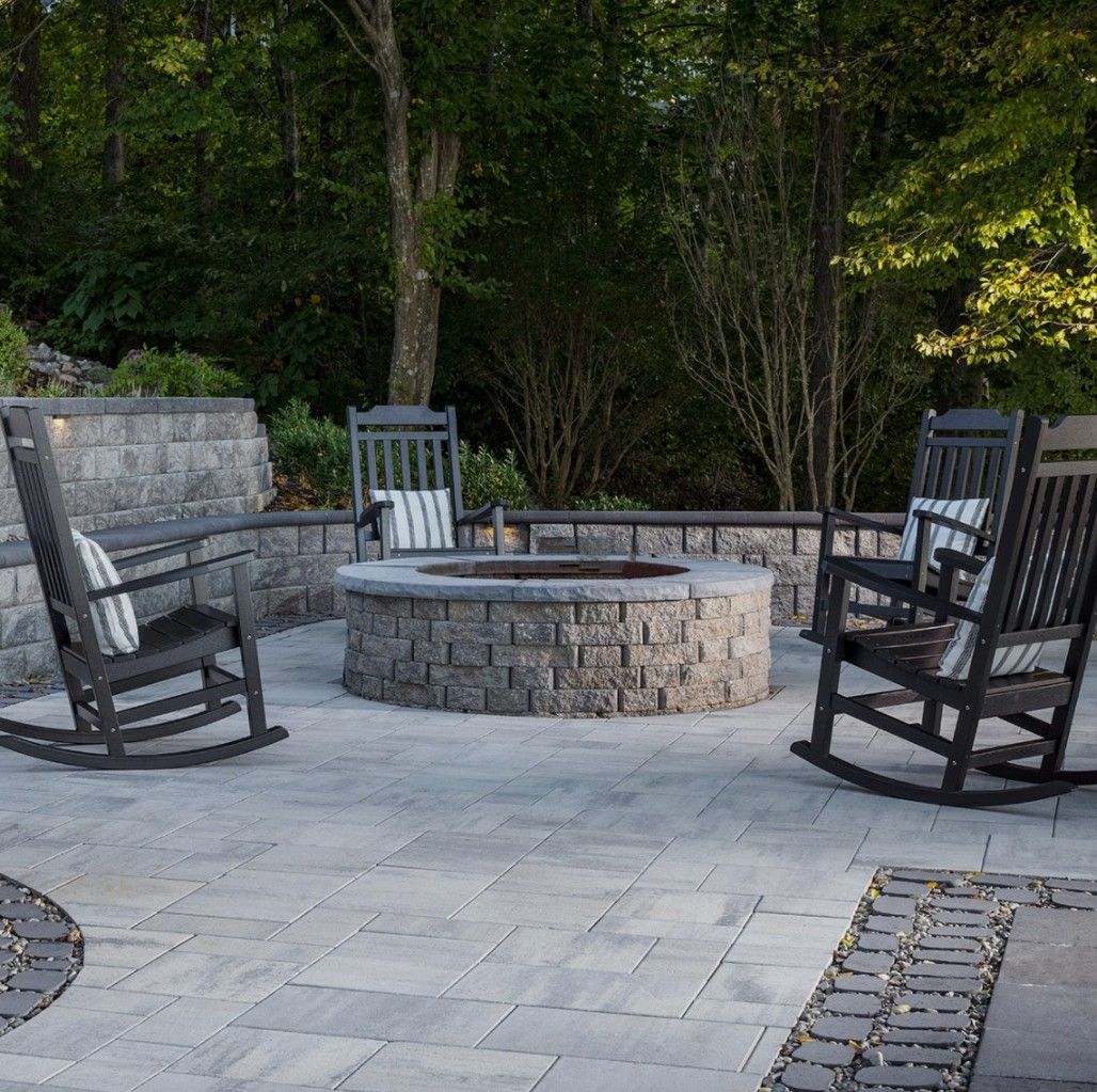 Stone fire pit with black rocking chairs on a paved patio surrounded by trees