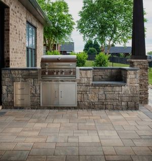 Outdoor stone patio with built-in stainless steel grill and cabinets beside a house, backed by trees and fencing