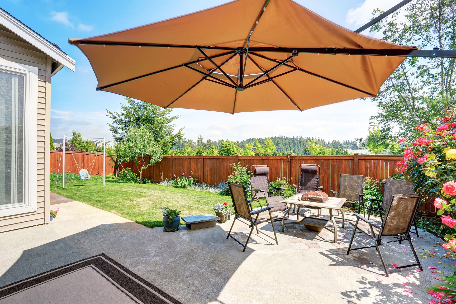 Backyard patio with large orange umbrella, outdoor seating, grill, and flower beds beside a house