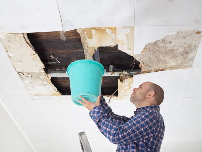 Man on ladder holding a turquoise bucket under a hole in a damaged ceiling.