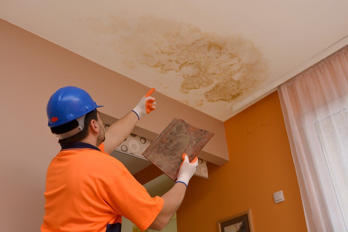 Worker in orange shirt and blue helmet examines water damage on a ceiling, holding a tile.