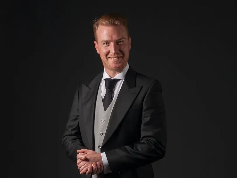 Een man in een smoking en stropdas staat met zijn handen gevouwen en glimlacht.