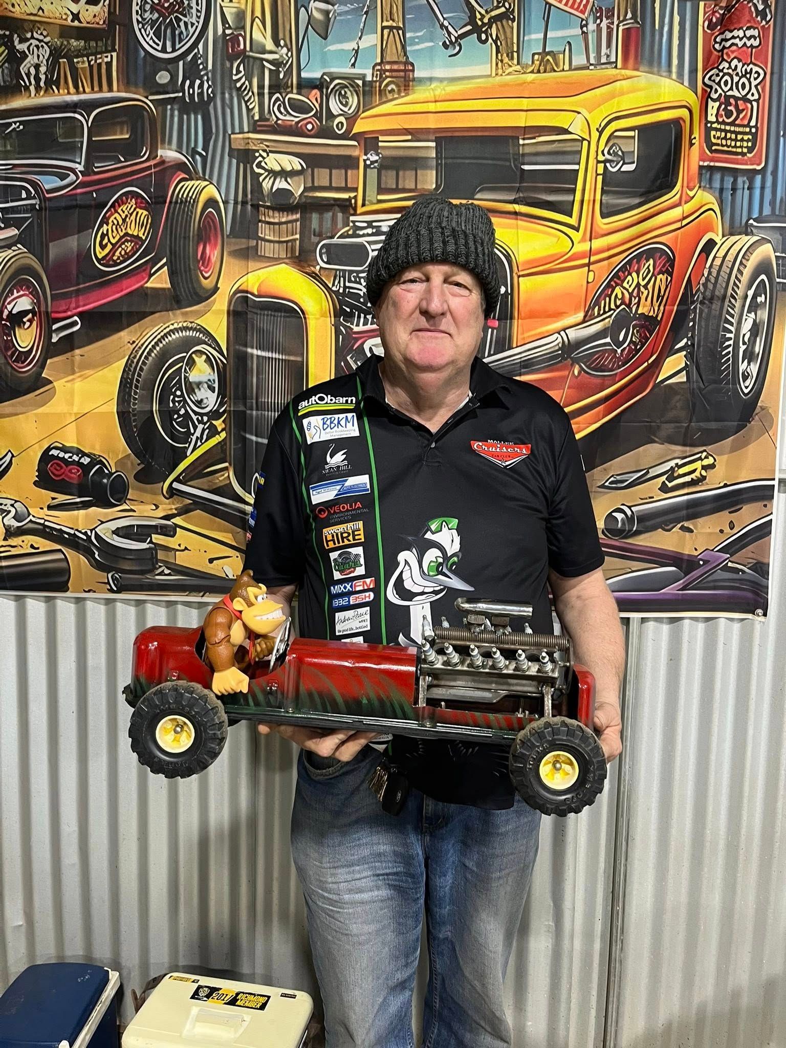Man standing in front of tin shed holding a small car 