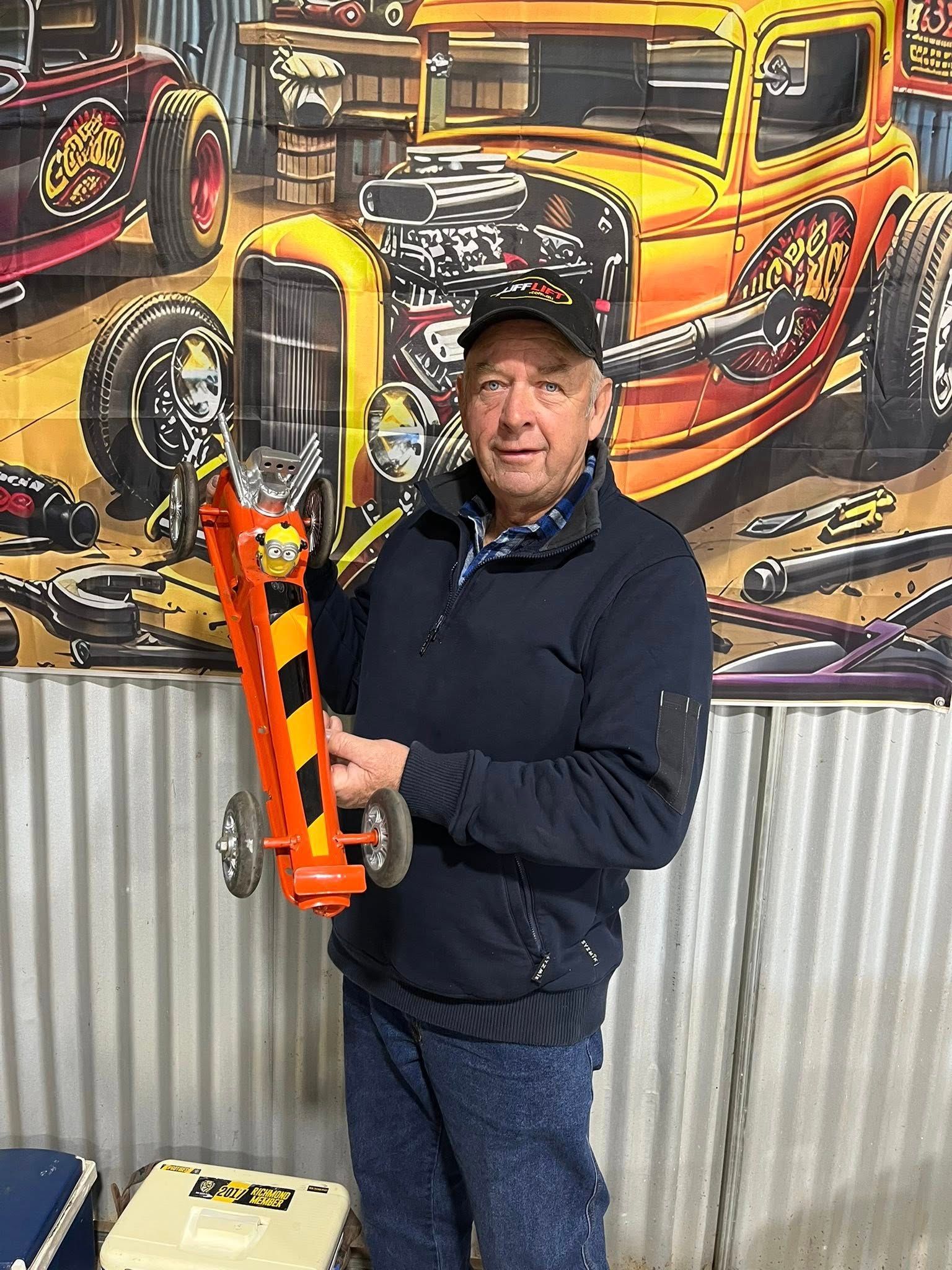 Man standing in front of tin shed holding a small car 