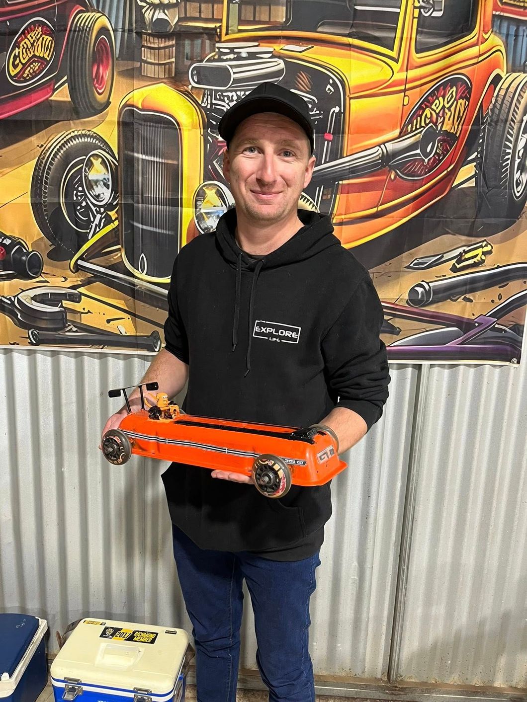 Man standing in front of tin shed holding a small car 
