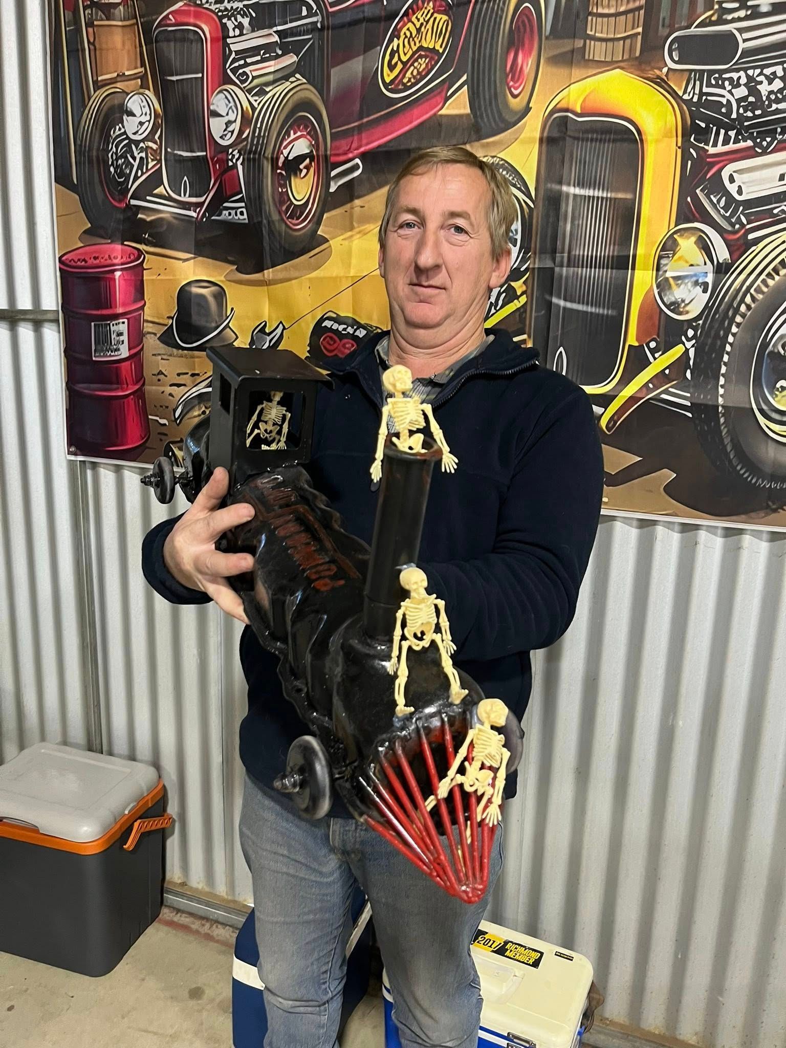 Man standing in front of tin shed holding a small car 