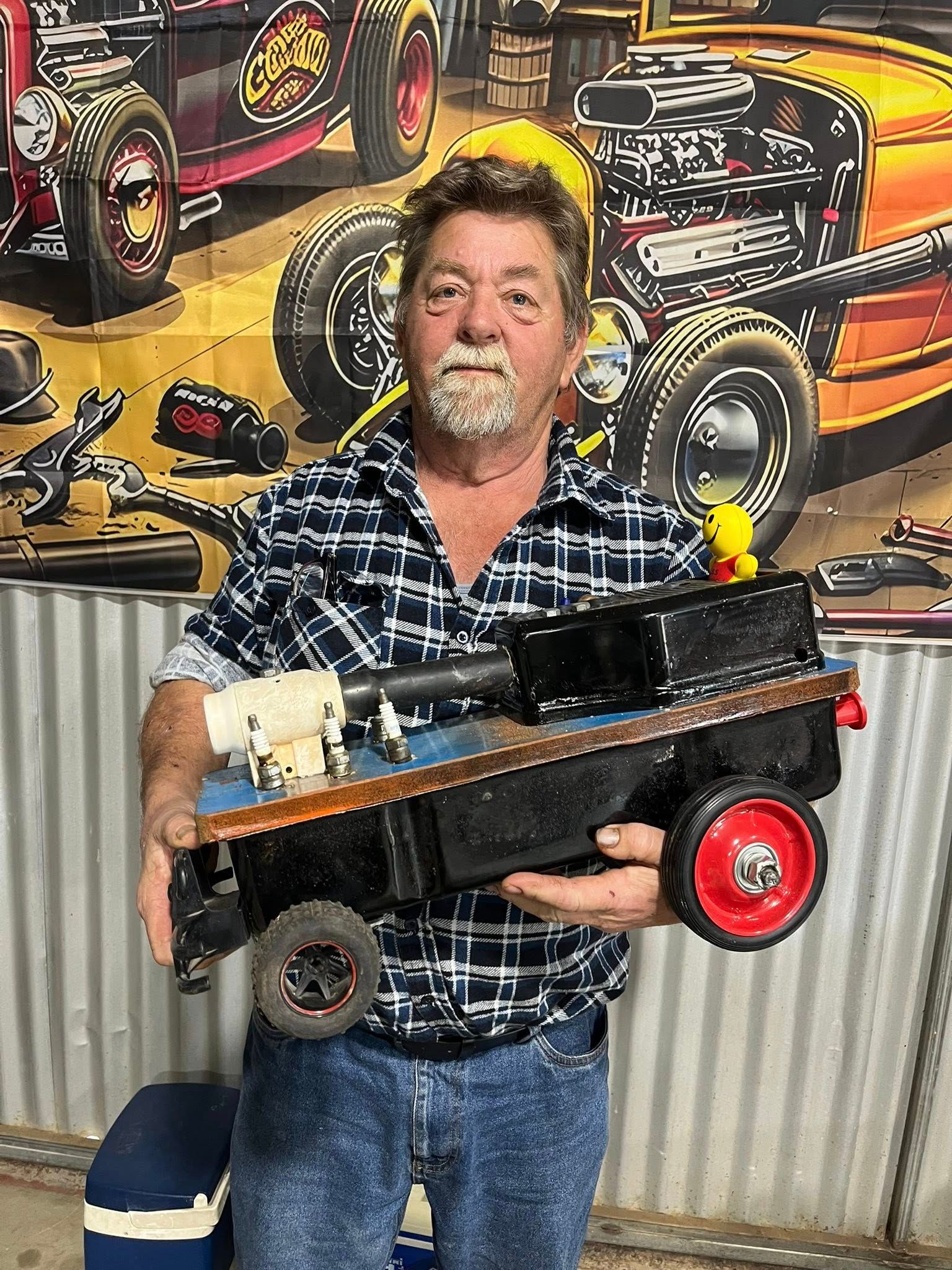 Man standing in front of tin shed holding a small car 