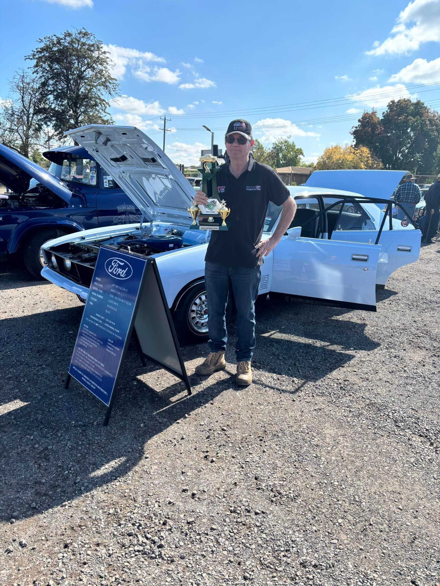 man stands in front of his car holding a trophy