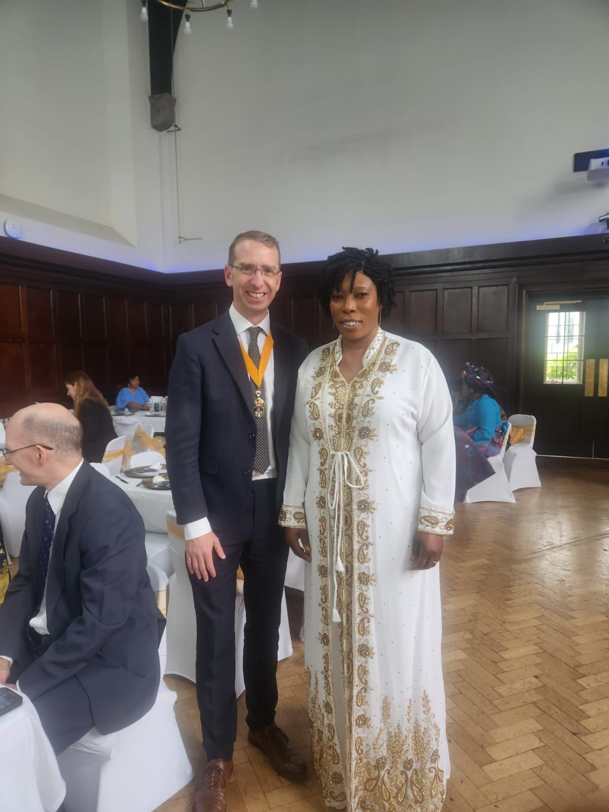A man in a suit and a woman in a white robe with gold trim stand indoors. They are smiling, possibly at an event.